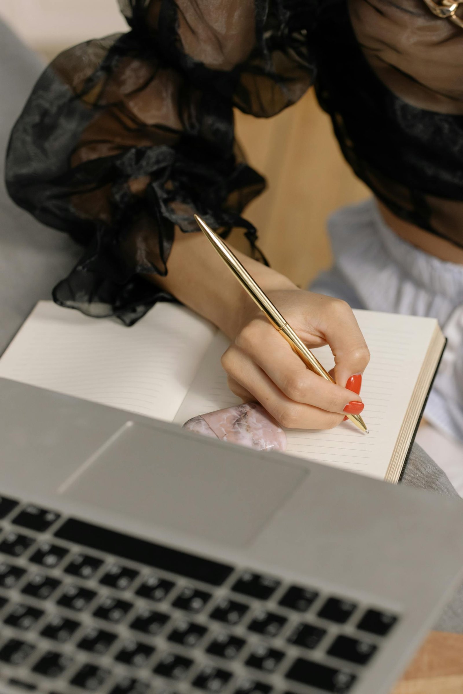 Woman in sheer black blouse writing in a notepad with a gold pen near a laptop.